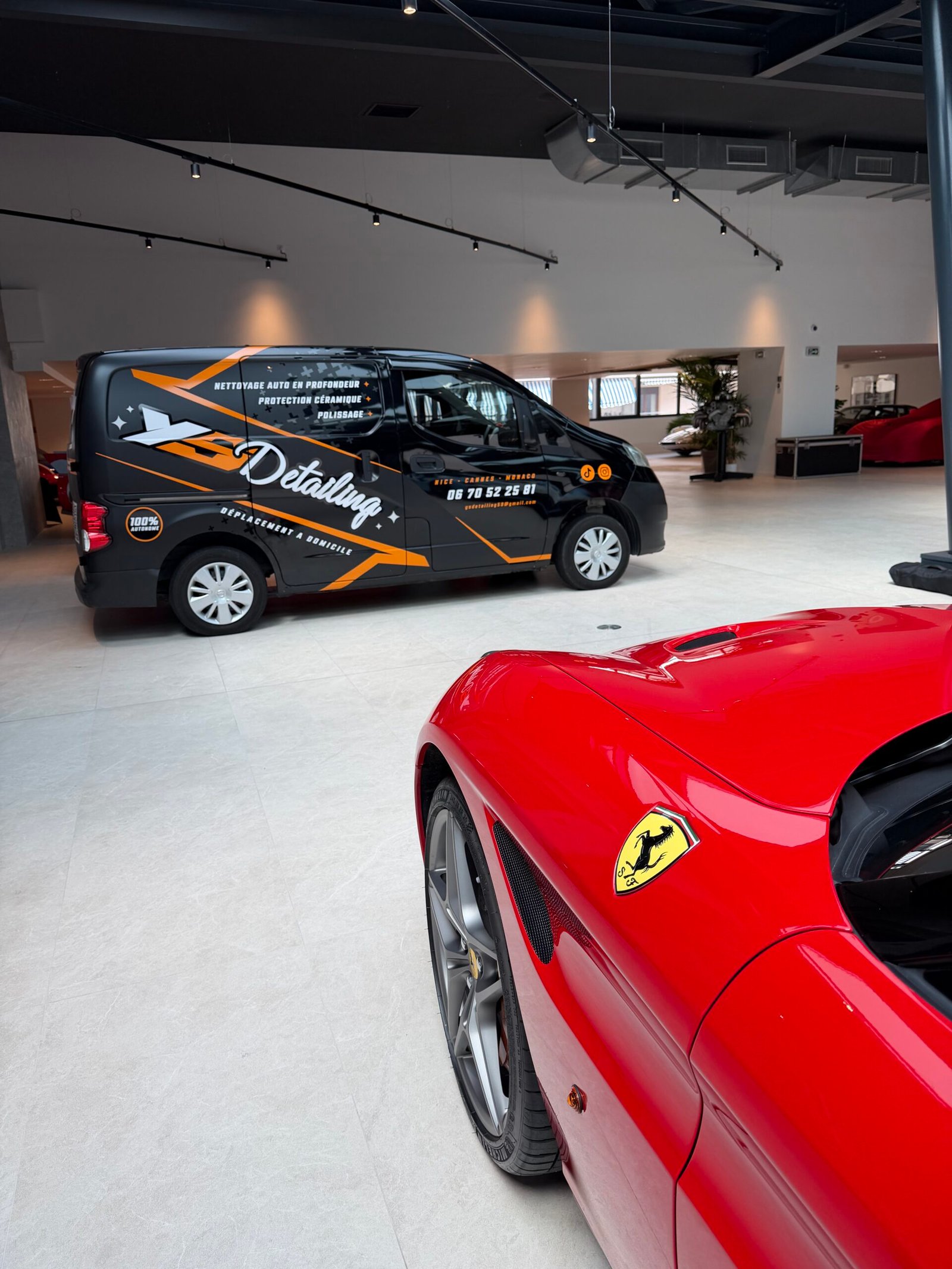 Ferrari rouge vue de trois-quarts avant dans le showroom YS Detailing, écusson Ferrari en premier plan, fourgon d'intervention en arrière-plan