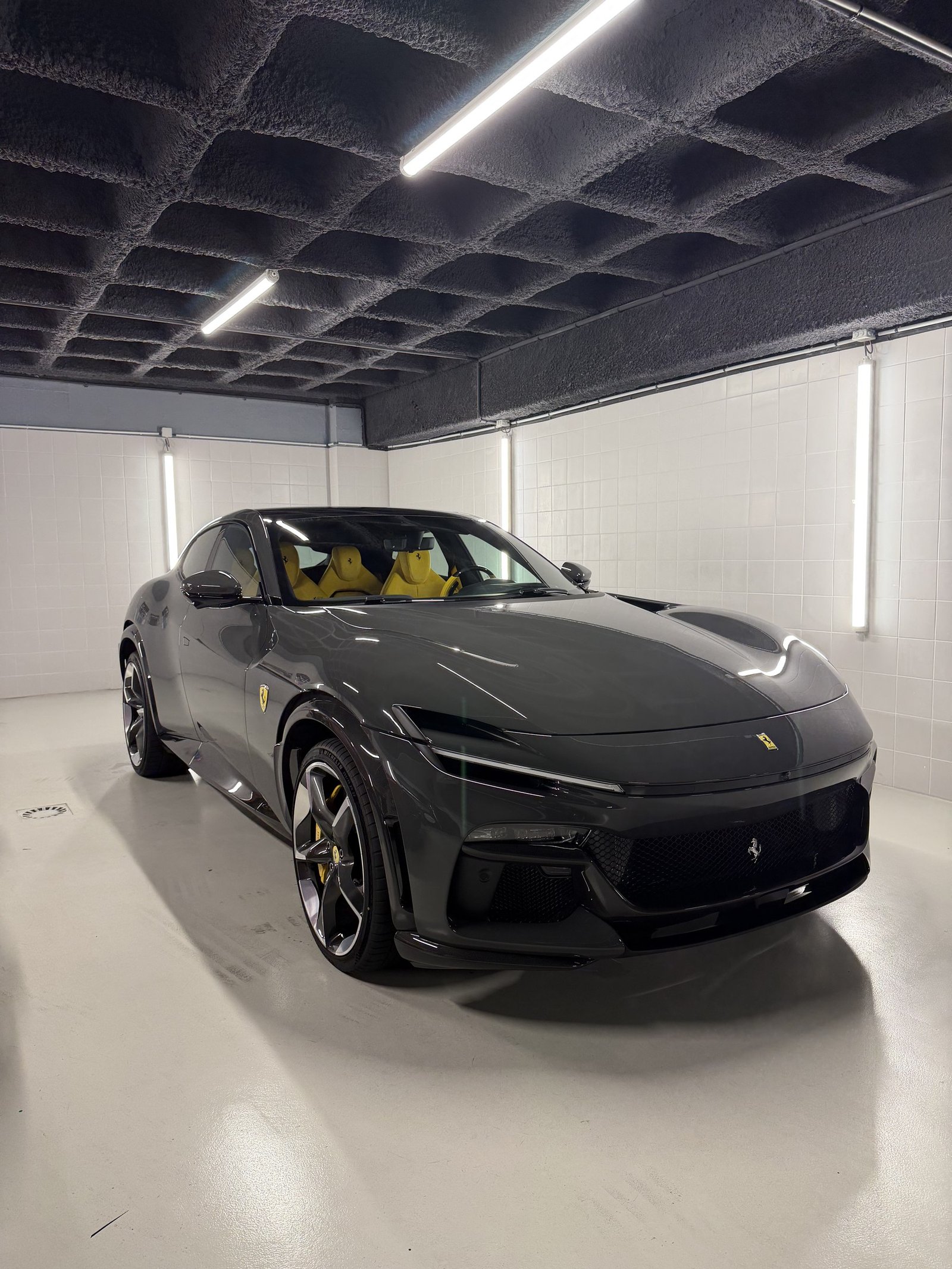 Ferrari noire vue de trois-quarts avant dans un box de detailing aux murs carrelés blancs, sellerie intérieure jaune visible
