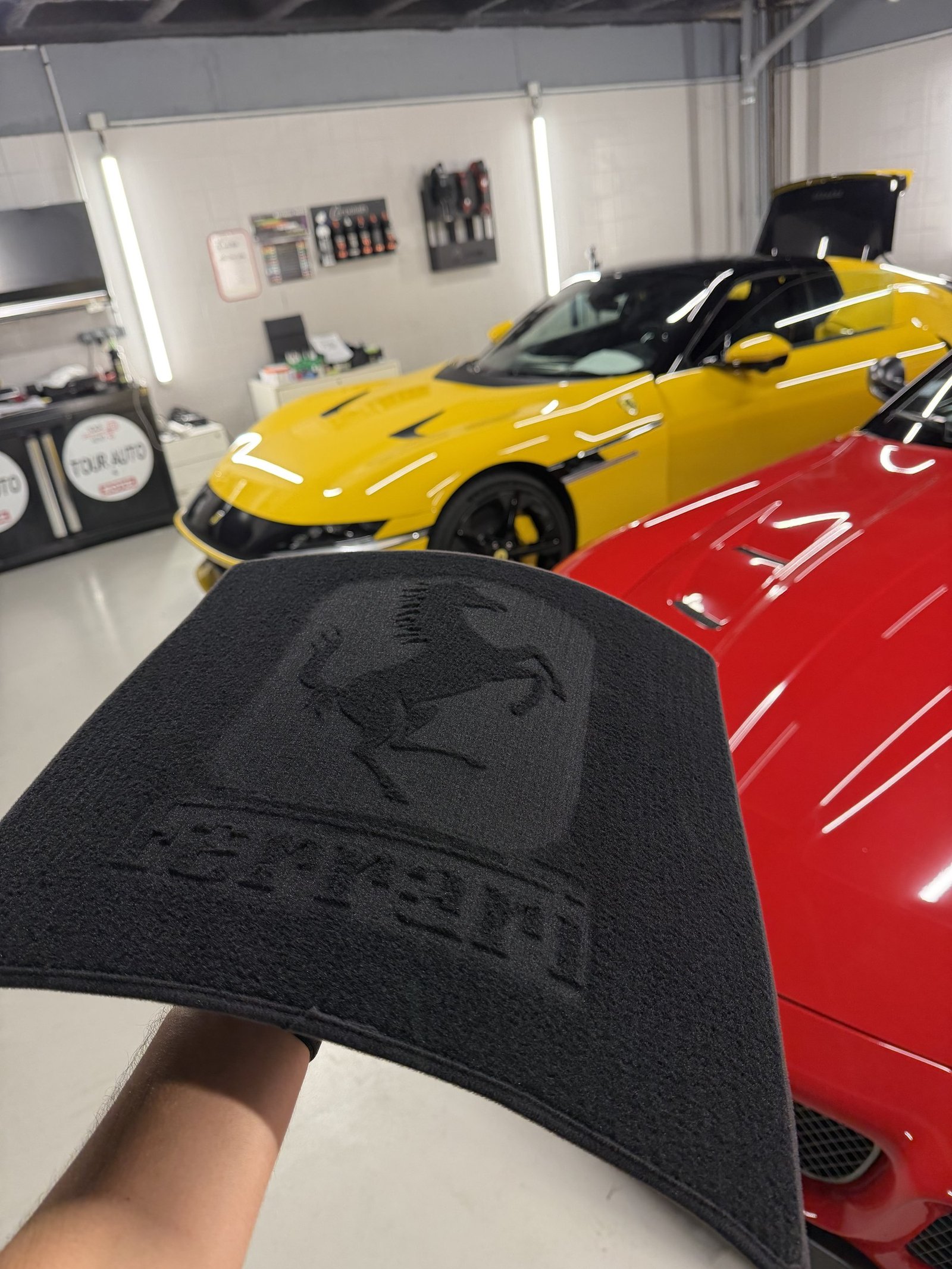 Tapis de sol floqué du blason Ferrari posé au premier plan dans l'atelier YS Detailing, avec une voiture jaune et une carrosserie rouge visibles en arrière-plan